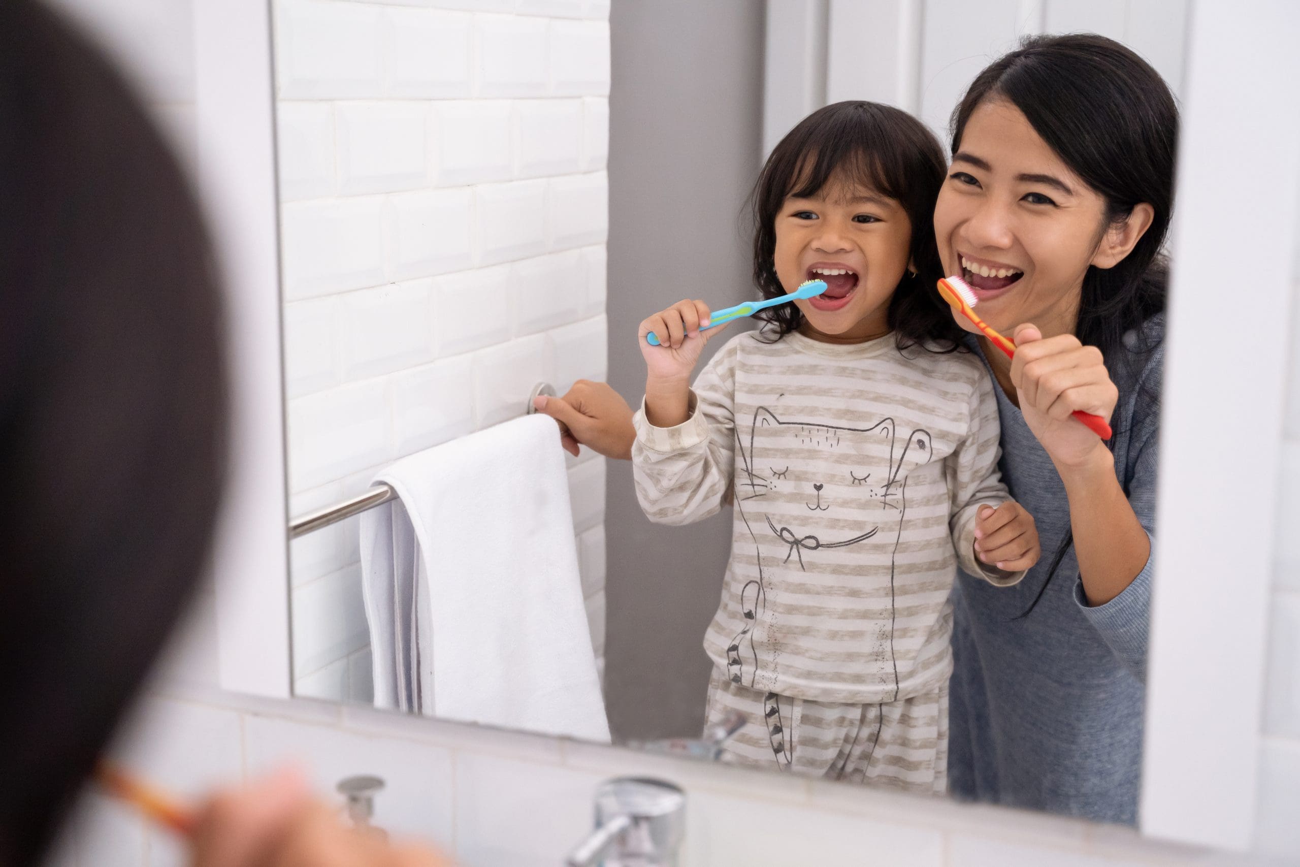 Mother and daughter brush their teeth in front of a mirror.