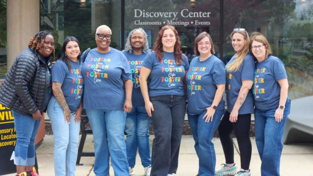 A group of eight diverse women standing side by side outdoors in front of a building labeled "Discovery Center." Most are wearing matching blue t-shirts printed with colorful text featuring the words "Foster Love," "Foster Hope," and "Foster Care." Everyone is smiling at the camera.